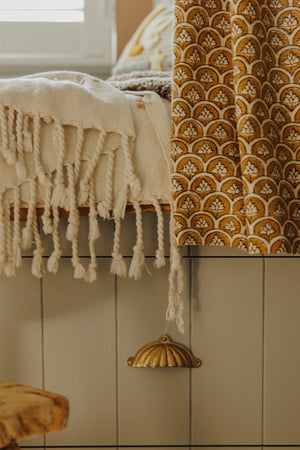 A close-up of a home interior showing a under bed built-in storage drawer with an aged solid brass scalloped cup handle. A cream knit throw blanket with tassels is draped over, next to a mustard yellow and cream patterned curtain. 