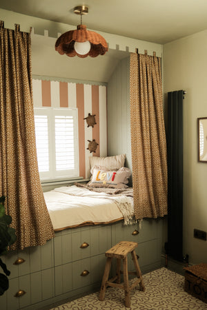 A cozy bedroom corner featuring a built-in daybed with aged brass cup handles on the storage drawers underneath. Patterned brown curtains, striped wallpaper, a small wooden stool, and a ceiling light fixture feature in the room.